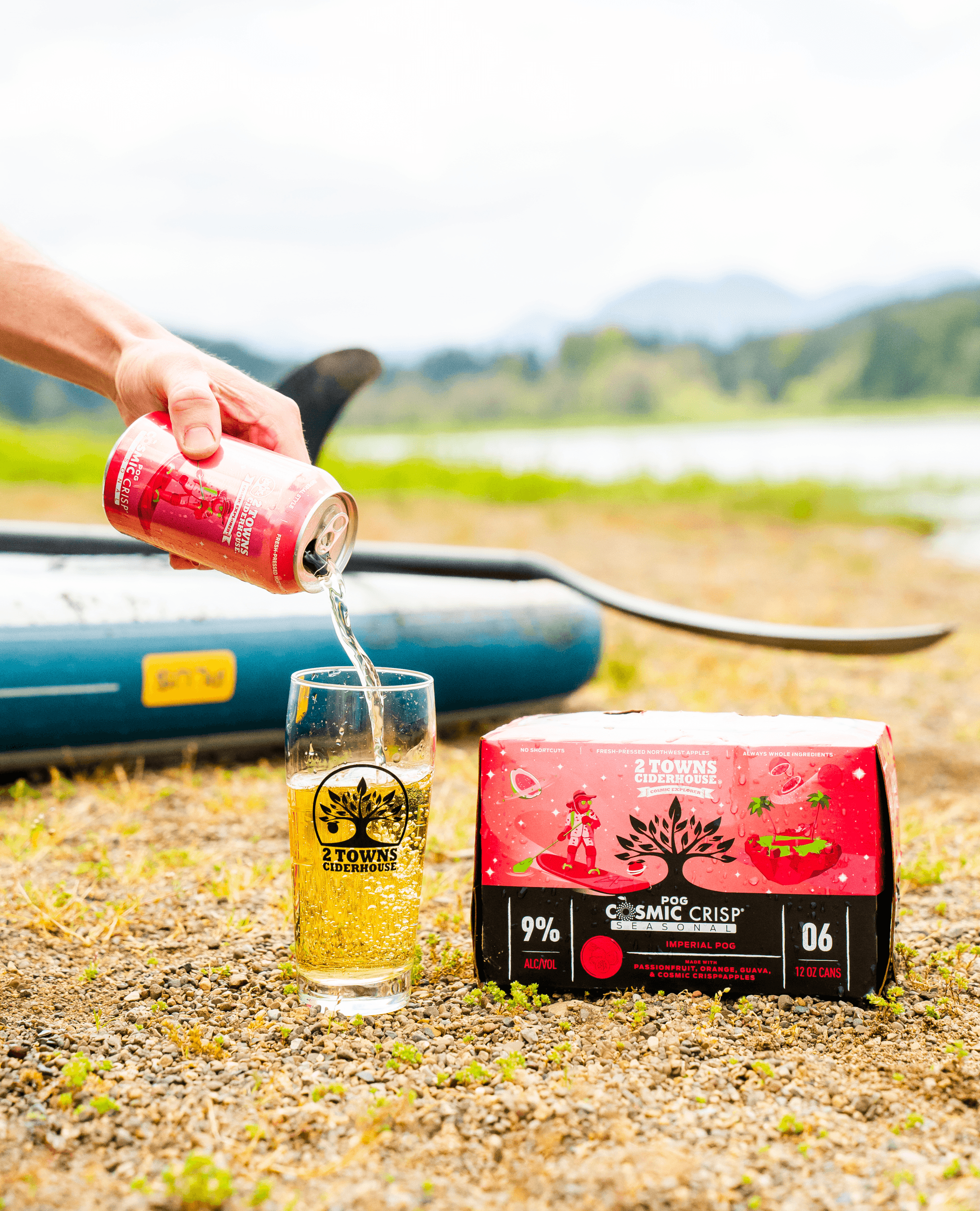 A hand pours a 2 Towns Cideerhouse POG Cosmic Crisp can into a glass on a rocky lakeshore. Next to the glass there is a 6-pack box of the same cider. A stand up paddleboard is in the background.